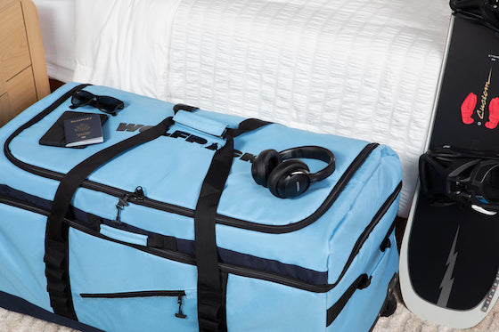 Blue WOLFPACK travel bag packed in hotel room with headphones, passport, and sunglasses on top next to snowboard